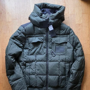 Museum grön dunjacka, storlek 52 Ny oanvänd  - Grön vadderad dunjacka med huva från Museum. Jackan har två fickor med lock framtill samt en dragkedja fram. Perfekt för kyligare dagar.  Ny med taggar Grön  Storlek 52.