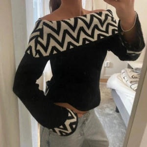 Svart stickad offshoulder-tröja - Säljer en svart stickad tröja med offshoulder-modell och bred vit kant med svart sicksackmönster över axlar och ärmslut. Tröjan har lång ärm och en lite loose passform, perfekt för dig som vill sticka ut med en cool och unik look.