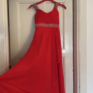 Röd långklänning med pärldetaljer - Snygg röd långklänning från Ami Kids Fashion med breda axelband och pärldetaljer på både axelband och midja. Klänningen har ett plisserat liv och ett flytande, svepande tyg som ger en elegant look. Perfekt för dig som vill sticka ut med färg och detaljer.