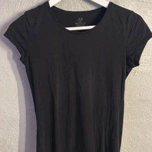 Svart basic t-shirt från H&M - En stilren svart t-shirt från H&M Basic, tillverkad i mjuk ekologisk bomull. Modellen har rund hals och korta ärmar, perfekt för en enkel och clean look. Passformen är normal och tyget känns skönt mot huden.