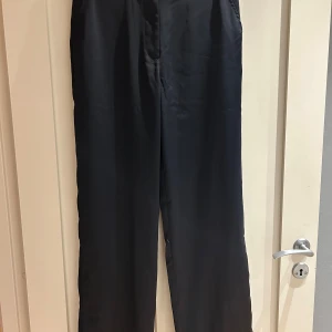 Svarta satin byxor från Bik Bok - Snygga svarta satin byxor från Bik Bok i storlek 38. Byxorna har raka ben, bälteshällor och sidfickor fram samt två fickor bak. Materialet har en lätt glansig finish som ger en stilren look. Perfekta för dig som gillar klassisk och clean stil.