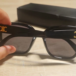 Svarta solglasögon med gulddetaljer - Säljer ett par svarta solglasögon med breda bågar och coola guldfärgade logodetaljer på sidorna. Glasen är mörka och bågarna har en fyrkantig form som ger en trendig vibe. Perfekt accessoar för att lyfta din stil.