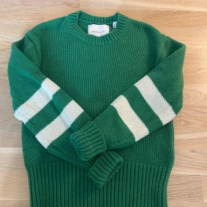 Grön stickad tröja med ränder från GANT - Snygg grön stickad tröja från GANT i storlek XS. Tröjan har vita ränder på ärmarna och en bred ribbad kant vid midjan och ärmsluten. Rund hals och avslappnad passform, perfekt för dig som gillar sportig och chill stil. 