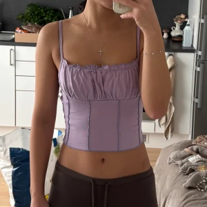 Ljuslila croppad korsett-topp - Säljer en ljuslila croppad korsett-topp med smala axelband och rynkad detalj vid bysten. Toppen har synliga sömmar framtill som ger en cool look och är tillverkad i mjukt bomullsmaterial. Perfekt för dig som gillar trendiga och feminina plagg.