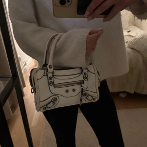 Väska - Säljer en vit handväska i skinn med coola metalldetaljer och dragkedja framtill. Väskan har två handtag och snygga svarta kontrastsömmar som ger den en edgy vibe. Perfekt storlek för det viktigaste och riktigt trendig design. Långt band tillkommer
