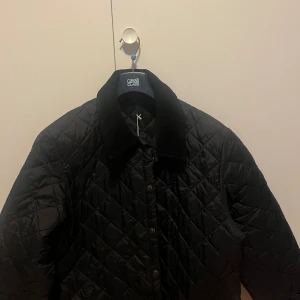 Svart quiltad jacka från Barbour - Quiltad svart jacka från Barbour i storlek L. Jackan har en klassisk krage i manchester, knappstängning framtill och ett stilrent rutmönster över hela ytan. Perfekt för dig som gillar tidlös och clean stil.