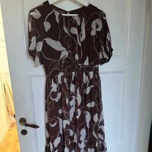 Brunvit blommig långklänning Stockh LM - Snygg långklänning från Stockh LM i brun och vit med blommigt mönster. Klänningen har korta puffärmar, markerad midja och volang nertill. Luftigt material som ger ett flowy intryck. Perfekt för dig som gillar en feminin och trendig stil.