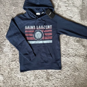 Saint Laurent Hoodie - Elegant hoodie från Saint Laurent i marinblått – prydd med vintageinspirerat tryck och ikonisk logga. En exklusiv statement-piece som kombinerar komfort med lyxig design.  ♦️ Storlek S – slim passform ⚜️ Helt nytt skick med tagg ♦️ Premiumkvalitet i 100% bomull  📦 Fraktas inom 24 timmar & trygg affär