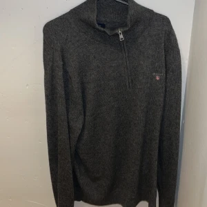 Mörkgrå stickad tröja från GANT - Mörkgrå stickad tröja från GANT med halv dragkedja vid halsen och broderad logga på bröstet. Tröjan har långärmad passform och är perfekt för kyliga dagar. Stilren och enkel design som funkar till jeans eller chinos.