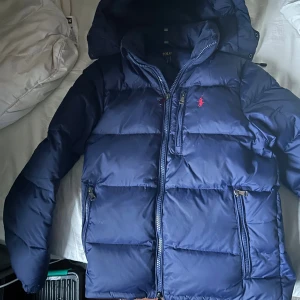 Blå pufferjacka från Polo Ralph Lauren - Snygg marinblå pufferjacka från Polo Ralph Lauren med huva och dragkedja framtill. Jackan har flera fickor med dragkedja och den klassiska röda Polo-loggan på bröstet. Perfekt för kalla dagar och har en riktigt schysst, puffig look. Kan gå ner i pris vid snabb affär.