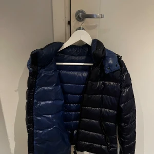 Mörkblå Moncler pufferjacka - Säljer en snygg mörkblå pufferjacka från Moncler med glansig finish och quiltad design. Jackan har huva med blå detaljer, två dragkedjefickor och Moncler-logga på insidan. Perfekt för kalla dagar och har en modern, sportig look. Köpt för 3 år sen via Plick, hon hade dinglat kvitto från moncler men har tyvärr inte kvar det. Storlek 2 vilket motsvarar S 