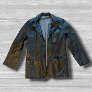 Svart skinnkavaj från Prestige - Svart kavaj i äkta skinn från Prestige, Collection de France. Klassisk modell med två knappar framtill, tre fickor och snygg krage. Fodrad insida i grått tyg. Perfekt för dig som vill ha en cool och tidlös look.