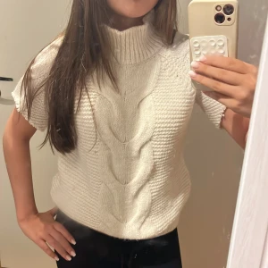 Trekvartsärmad polotröja - Säljer en vit stickad polotröja med korta ärmar och grov kabelstickning framtill. Tröjan har en hög ribbad krage och är gjord i mjukt material som känns riktigt cozy. Perfekt för lager-på-lager och chill stil.armarna är omgjorda till kortare/trekvsrtsärm 