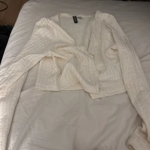 Vit omlottopp från H&M Divided - Säljer en vit omlottopp från H&M Divided i storlek M. Toppen har lång ärm och ett quiltat mönster som ger en cool textur. Croppad passform och omlott-design framtill gör den extra trendig. Perfekt att styla med höga jeans eller kjol.