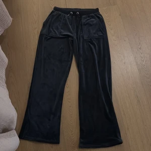 Mörkblå mjukisbyxor Juicy Couture - Svarta mjukisbyxor från Juicy Couture med breda ben och snörning i midjan. Byxorna har stora fickor fram och bak med knappdetaljer, samt broderad JC-logga på ena fickan. Mjukt och glansigt material som känns lyxigt. Och har andast använt minst 2 gånger. 