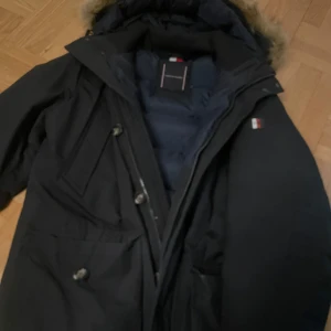 Svart parkas från Tommy Hilfiger L - Svart parkas från Tommy Hilfiger i storlek L med pälskantad huva och klassisk TH-logga på ärmen. Jackan har flera fickor med knappar och dragkedja framtill. Fodret är quiltat och jackan är perfekt för kalla dagar.