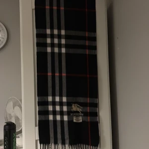 Svart rutig halsduk från Burberry - Snygg svart halsduk från Burberry med klassiskt rutigt mönster i vitt, grått och röda linjer. Halsduken har fransar i ändarna och en broderad Burberry-logga. Perfekt accessoar för att lyfta din outfit med lite lyxig vibe.