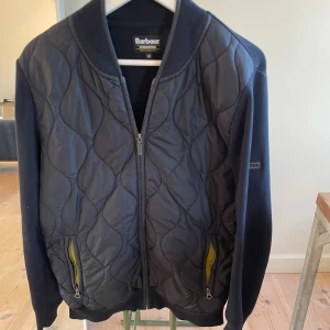 Quiltad bomberjacka från Barbour - Snygg mörkblå bomberjacka från Barbour International med quiltad framsida och stickade ärmar och rygg. Jackan har dragkedja framtill, två fickor med gul insida och ribbad krage. Perfekt för dig som gillar stilren och sportig look. Storlek M