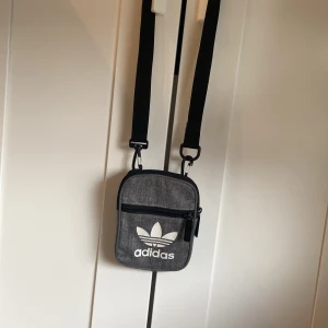 Grå axelväska från Adidas - Snygg grå axelväska från Adidas med klassisk vit logga framtill. Väskan har justerbar svart rem, dragkedja och ett extra fack på framsidan. Hör av vid funderingar, pris kan diskuteras! 