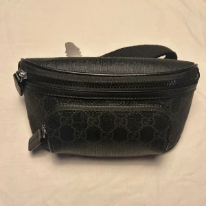Svart Gucci magväska med GG-mönster  - Snygg svart Gucci magväska  med klassiskt GG-mönster i mörkgrått. Väskan har tre fickor två fram och en i bak sidan. Det finns fickor inuti väskan, skriv för mer info..