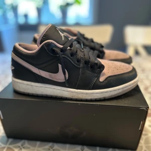 Nike Jordan 1 Low svart/rosa sneakers - Nike Jordan 1 Low sneakers med svart skinn och ljusrosa mockadetaljer på tå och Swoosh. Klassisk Jumpman-logga på plösen, svarta snören och vit platt sula. Snygg kontrast mellan färgerna och låg profil som passar streetwear-stilen.