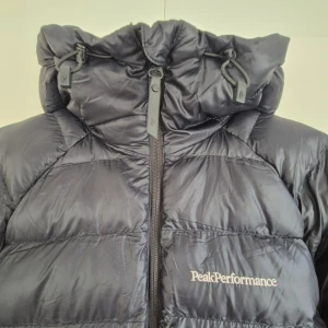 Svart Peak Performance dunjacka - Jacka somär perfekt för den stundande vintern, snygg med detaljer på huva och märke fram. fylld med syntetfri fiberisolering som efterliknar dun och håller dig varm även i våta förhållanden. Passform: Regular. Puffad modell.  Köpt ifjol, lite använd och i nyskick.  Nypris 3.200:-