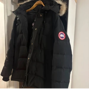 Svart dunjacka från Canada Goose - Svart dunjacka från Canada Goose med stor huva och fluffig pälsdetalj. Jackan har det klassiska runda Canada Goose-märket på ärmen, dragkedja och knappar framtill. Quiltad design och långärmad modell, perfekt för kalla vinterdagar. Storlek L men passar M.