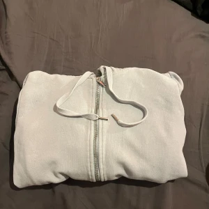 Vit hoodie med dragkedja och huva - Säljer en stilren vit hoodie med dragkedja framtill och snörning i huvan. Perfekt för chill dagar eller när du vill ha en enkel och clean look. Mjuk och skön, passar till det mesta i garderoben.