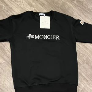 Svart sweatshirt från Moncler - Svart sweatshirt från Moncler med vit broderad logga på bröstet och klassisk rund hals. Tröjan har Moncler-märke på ena ärmen och är tillverkad i mjuk bomull. Perfekt för dig som gillar stilrena och exklusiva plagg.