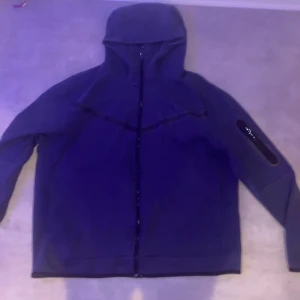 Nike tech set  - Nike hoodiejacka i mörkblå färg med hel dragkedja och huva. Jackan har långärmad design och en ficka med dragkedja på ena ärmen. Tillverkad i mjukt bomullsmaterial, perfekt för chill och streetwear. Snygg och enkel stil med diskret Nike-logga.