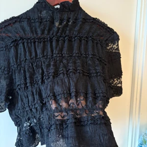 Svart spetsblus med volanger - Svart blus med korta ärmar och hög hals. Blusen är gjord i spets med flera lager av volanger och har en transparent look som ger en cool vibe. Perfekt för dig som gillar detaljer och vill sticka ut med en unik topp.  Endast använd en gång, och fick många komplimanger!! Passar mig som vanligtvis bär L/M då den är stretchig men magen syns lite om man bär lowwaist jeans. Passar för dig som är strl S för mer vanlig passform / kort överkropp 