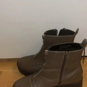 Bruna boots med dragkedja - Snygga bruna boots i skinn med grov platt sula och silverfärgad dragkedja framtill. Modellen har rund tå och markerade sömmar som ger en cool look. Perfekt för dig som gillar stilrena och robusta skor med edge.