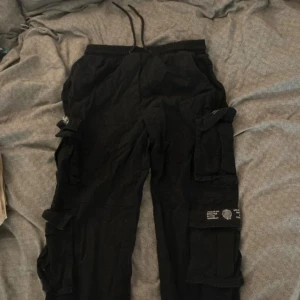 Svarta cargopants från FB Sister - Svarta cargopants från FB Sister med elastisk midja och snörning. Flera stora fickor på benen, varav en med coolt tryck och grafisk detalj. Byxorna har ribbade muddar vid bensluten. Perfekt för en avslappnad streetstil.