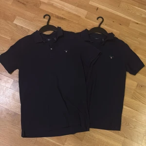 Marinblå pikétröjor från GANT - Två stilrena marinblå pikétröjor från GANT med klassisk krage och knappar framtill. Tröjorna har GANT-logga broderad på bröstet och är gjorda i mjuk bomull som känns skön mot huden. Perfekt för dig som gillar en clean och sportig look. Säljer dom för sig