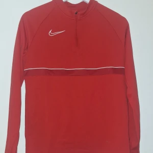 Röd Nike Dri-Fit långärmad tröja - Nike Dri-Fit långärmad tröja i rött med vit logga och tunn vit linje över bröstet. Tröjan har half zip framtill och är tillverkad i ett lätt, syntetiskt material som andas. Perfekt för träning och sportiga aktiviteter.