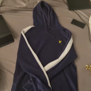 Marinblå hoodie från Lyle & Scott - Snygg marinblå hoodie från Lyle & Scott med vit rand längs ärmarna och klassisk gul logga på bröstet. Tröjan har huva med snörning och är tillverkad i mjuk bomull. Den har en liten skada vid magfickan