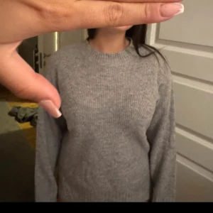 Grå stickad tröja med rund hals - Mysig grå stickad tröja med rund hals och ribbad struktur. Tröjan har långärmade ärmar och en klassisk, enkel design som är lätt att matcha med olika outfits. Perfekt för kyliga dagar och skön att dra på sig när man vill vara bekväm.