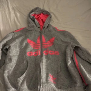 Grå och rosa Adidas hoodie XXL - Säljer en grå hoodie från Adidas i storlek XXL med rosa detaljer och logga framtill. Tröjan har huva med rosa insida, dragkedja och långa ärmar med rosa ränder längs axlarna. Perfekt för dig som gillar sportig och chill stil.