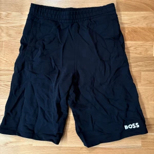 Svarta shorts från BOSS  - Svarta shorts från BOSS med enkel och clean design. De har elastisk midja, två sidofickor och BOSS-logga i vitt på ena benet. Tillverkade i mjuk bomullsblandning med lite polyester för extra komfort. Perfekta för chill dagar eller träning.