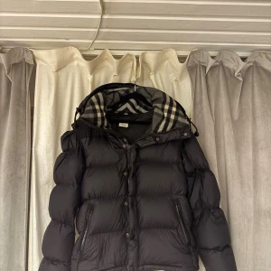 Svart Burberry pufferjacka med huva - Svart pufferjacka från Burberry med klassisk rutmönstrad huva. Jackan har dragkedja och knappar framtill, två fickor med dragkedja och en quiltad, puffig design. Materialet är polyester och dun/fjädrar, perfekt för kalla dagar. Snyggt Burberry-tyg i huvan ger extra stil.
