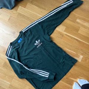 Mörkgrön långärmad Adidas tröja  - Säljer en mörkgrön långärmad tröja från Adidas med klassiska vita ränder längs ärmarna och Adidas logga på bröstet. Tröjan har rund hals och är i mjuk bomull, perfekt för en streetstyle-look. 