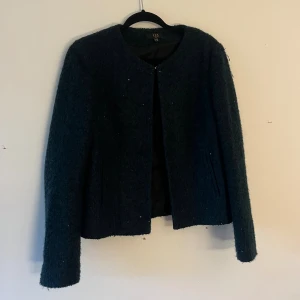 Mörkgrön ullkavaj från 1.2.3 - Snygg mörkgrön kavaj i ull från 1.2.3 med rund halsringning och lång ärm. Jackan har en stilren, rak passform och är helt utan knappar eller dragkedja framtill. Perfekt för dig som gillar en clean look och mjukt material.