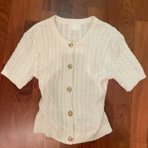 Vit kabelstickad kofta från H&M - Säljer en vit kabelstickad kofta från H&M i storlek S. Koftan har korta ärmar, rund halsringning och snygga guldfärgade knappar framtill. Ribbad kant vid ärmslut och nederkant ger en schysst passform. Perfekt för lager-på-lager under vår och höst.