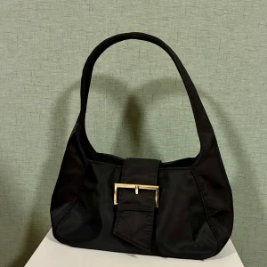 Svart axelväska från Bik Bok - Stilren svart axelväska från Bik Bok i syntetmaterial. Väskan har en bred axelrem och en snygg guldfärgad spänndetalj framtill. Perfekt storlek för det viktigaste och enkel att matcha med olika outfits.