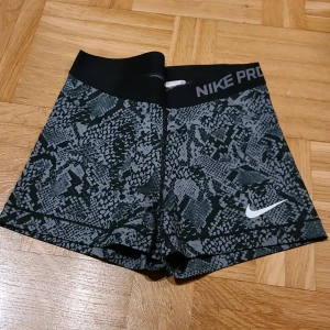 Nike Pro svarta träningsshorts S - Nike Pro shorts i svart med grått camouflage-mönster och bred elastisk midja. Snygg, tight passform som sitter skönt vid träning. Materialet är syntet och designen är sportig med Nike-logga på benet. Perfekta för gymmet eller löpning.