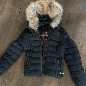 Svart pufferjacka med fuskpäls från Hollies - Svart pufferjacka från Hollies med fluffig fuskpäls på huvan och leopardmönstrat innerfoder. Jackan har guldfärgade dragkedjor, quiltad design och figurnära passform. Perfekt för kalla dagar med snygg streetstil.