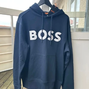 Ny Marinblå hoodie från BOSS med logga - Snygg marinblå hoodie från BOSS med stor vit logga framtill. Tröjan har huva med snörning och en klassisk känguruficka. Tillverkad i mjuk bomullsmix som känns skön mot huden. Perfekt för en chill och stilren look.med kapparna kvar