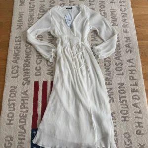 Vit omlottklänning från Gina Tricot - Supersnygg långklänning i vit från Gina Tricot med omlottmodell och v-ringning. Klänningen har långa ärmar med volangdetaljer vid ärmslut och ett knytband i midjan. Materialet är lätt plisserat och ger en cool struktur. Perfekt för dig som gillar stilrena och trendiga looks.