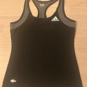 Svart och grå träningstopp från Adidas - Säljer en svart och grå linnetopp från Adidas med brottarrygg och Climacool-tryck. Toppen är i ett stretchigt material av polyester och spandex, perfekt för träning. Adidas-logga framtill och snygg kontrast mellan svart och grått tyg.