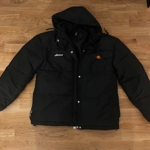Svart pufferjacka från Ellesse - Svart pufferjacka från Ellesse med huva och dragkedja framtill. Jackan har Ellesse-logga på bröstet och är vadderad för extra värme. Perfekt för kalla dagar och har en clean, sportig look.